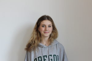 Hadley Marshburn, a teenage woman with brown hair fading into blond , smiling at the camera. She wears a grey sweatshirt and is standing in front of a white wall.