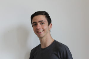 Jordan Galloway, a brunette teenage man smiling at the camera. He is wearing a green cotton t shirt and standing in front of a white wall. 