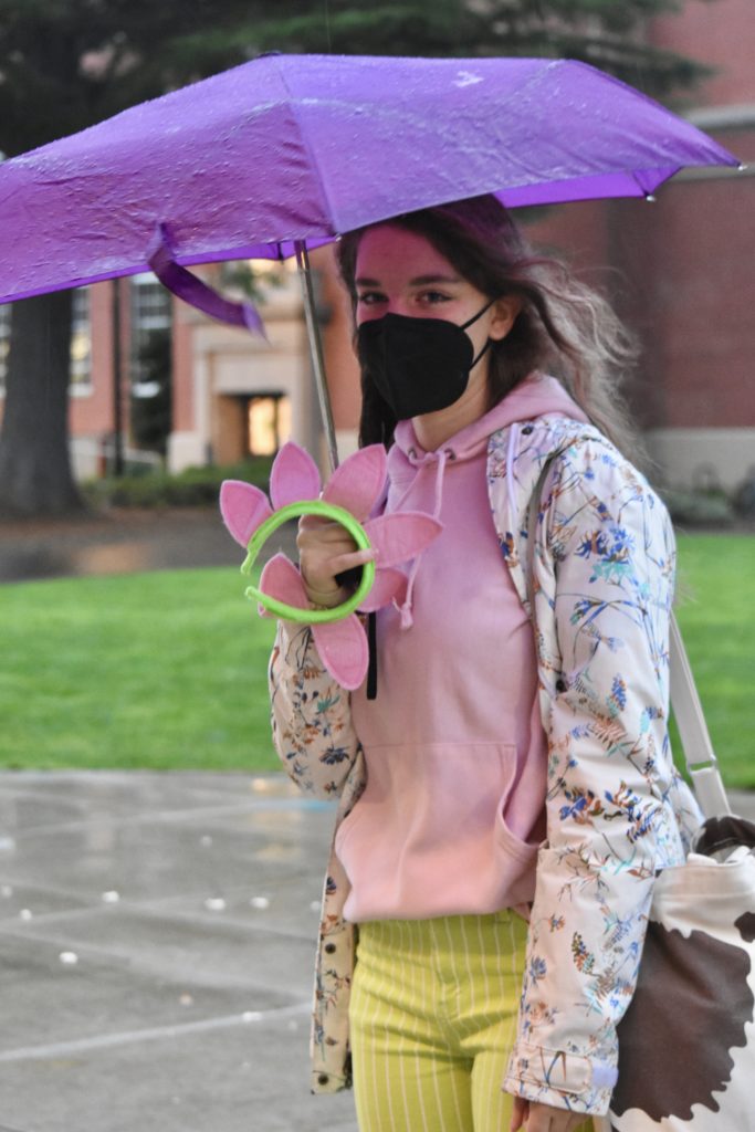 Miller stands outside Grant High School in the rain. They are wearing a pink sweat shirt, yellow pants, and a light patterned rain jacket. They are holding a pink flower pedal head band and a purple umbrella.