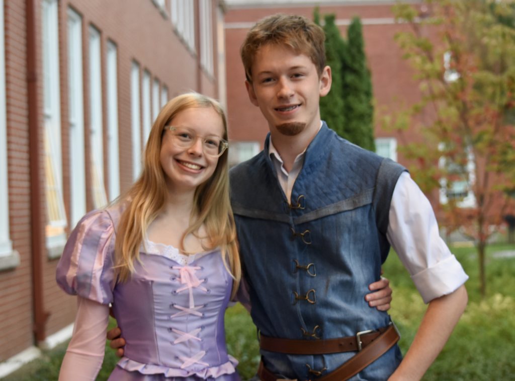 Loving and Hayden stand next to each other in front of a brick wall.  Loving is wearing a purple dress and Hayden is wearing a blue vest with a brown beard painted onto his chin. 