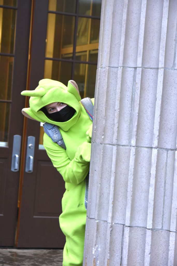 Baker, dressed in a neon green Teletubby suit, peeks out from behind a stone pillar. 