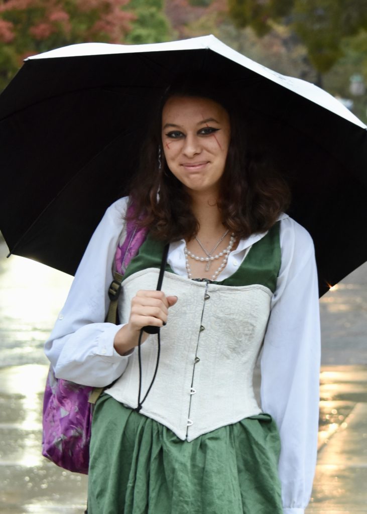 Korpi stands under a black umbrella wearing a green and white dress. A purple backpack hangs over her shoulder. 