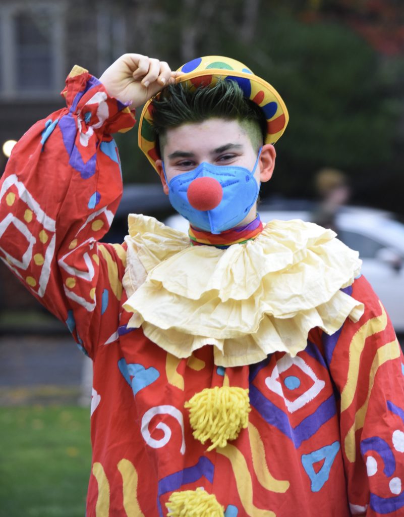 Mason wears a red, patterned clown suit with a ruffled collar and yellow pom-poms. Their hand reaches to tip their yellow polka-dotted hat, and a red clown nose is attached to their blue mask. 