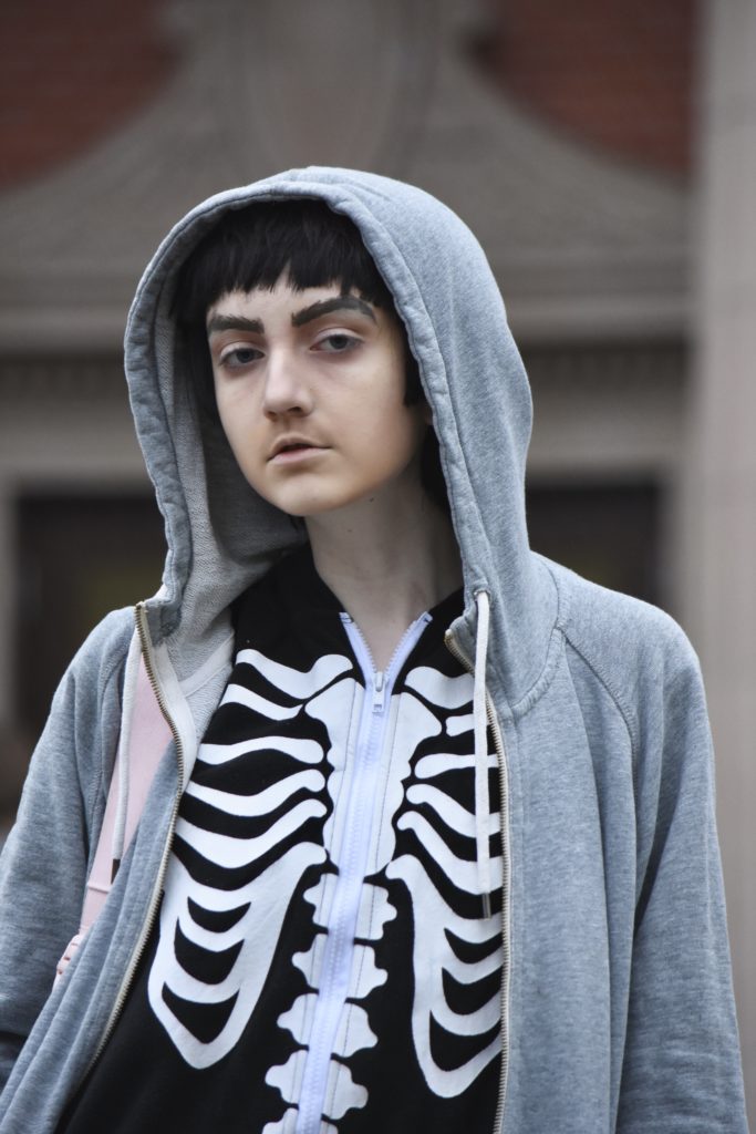 Hasseries wears a zipped-up skeleton suit and a gray sweatshirt with the hood up. 