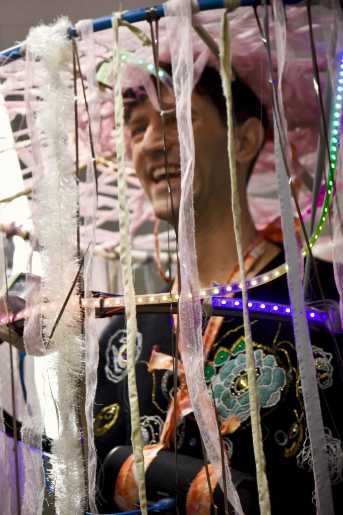 Mx. Lodore laughs behind a collection of shiny pink ribbons, which are attached to their head by a blue hula hoop. 