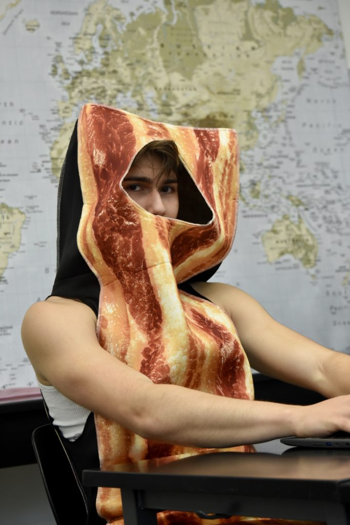 Peterson sits at a desk in front of a world map, his head peeking through a bacon costume. 