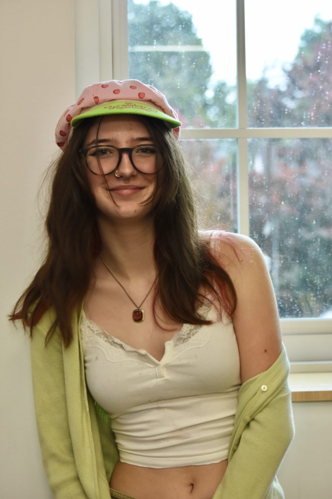 Rideout stands in front of a window, wearing a pink and green strawberry hat with a white shirt and green sweater. 