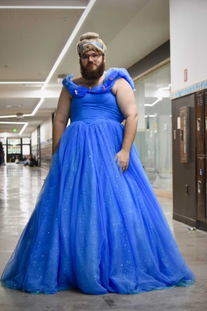 Mr. Kurzer stands in a hallway, wearing an elegant blue, sparkly dress and a wig of blonde hair in a bun. 
