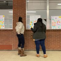 Two parents wearing fleece jackets point to a brick wall with a white poster covered in colorful sticky-notes.