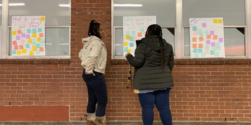 Two parents wearing fleece jackets point to a brick wall with a white poster covered in colorful sticky-notes.