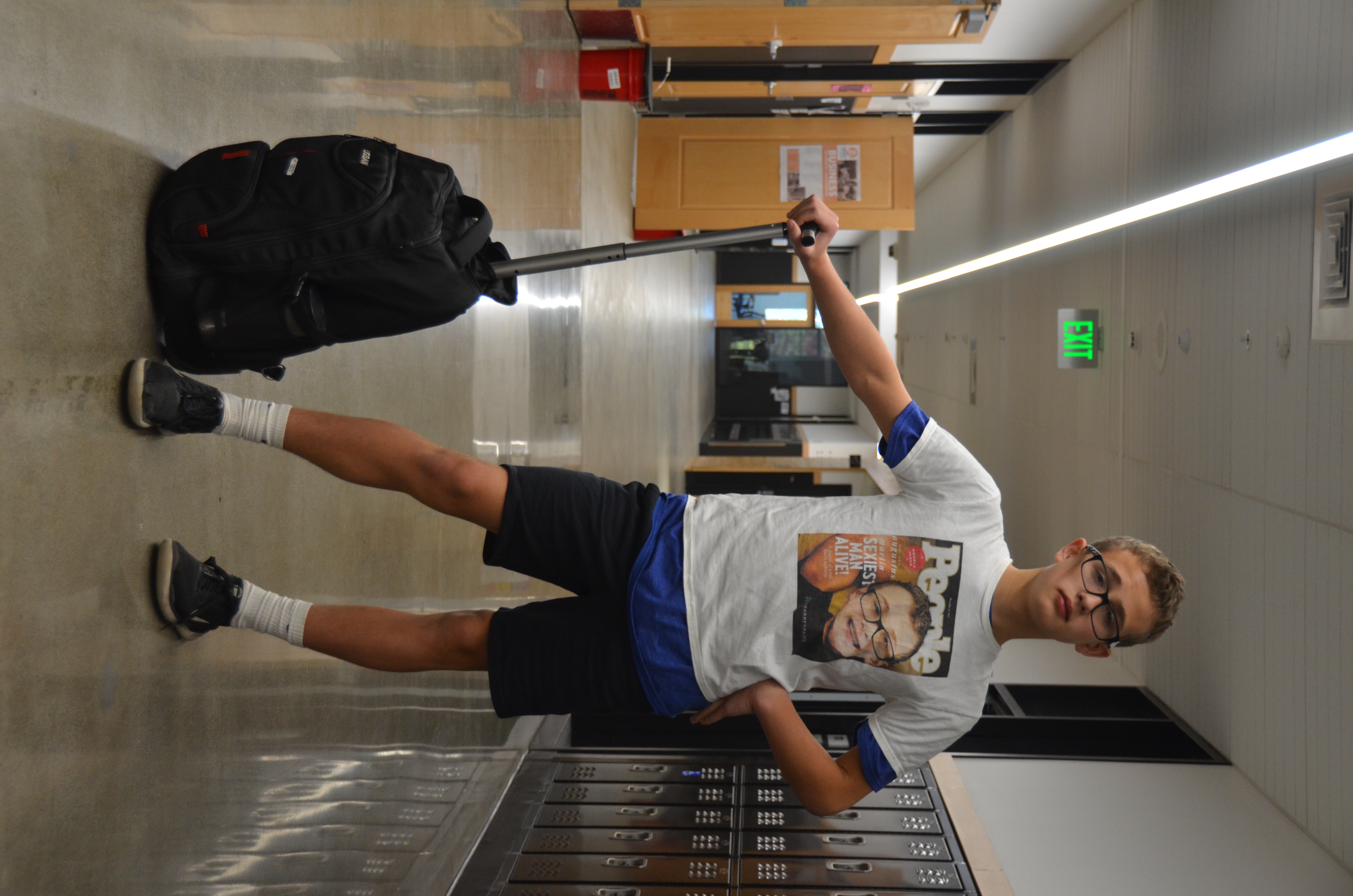 A boy with glasses and short blond hair stands in a school hallway with his rolling backpack