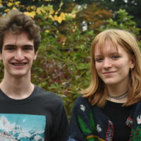Two white teenagers, one with short brown hair and another with strawberry blonde hair and bangs, stand in front of a wall of greenery, smiling.