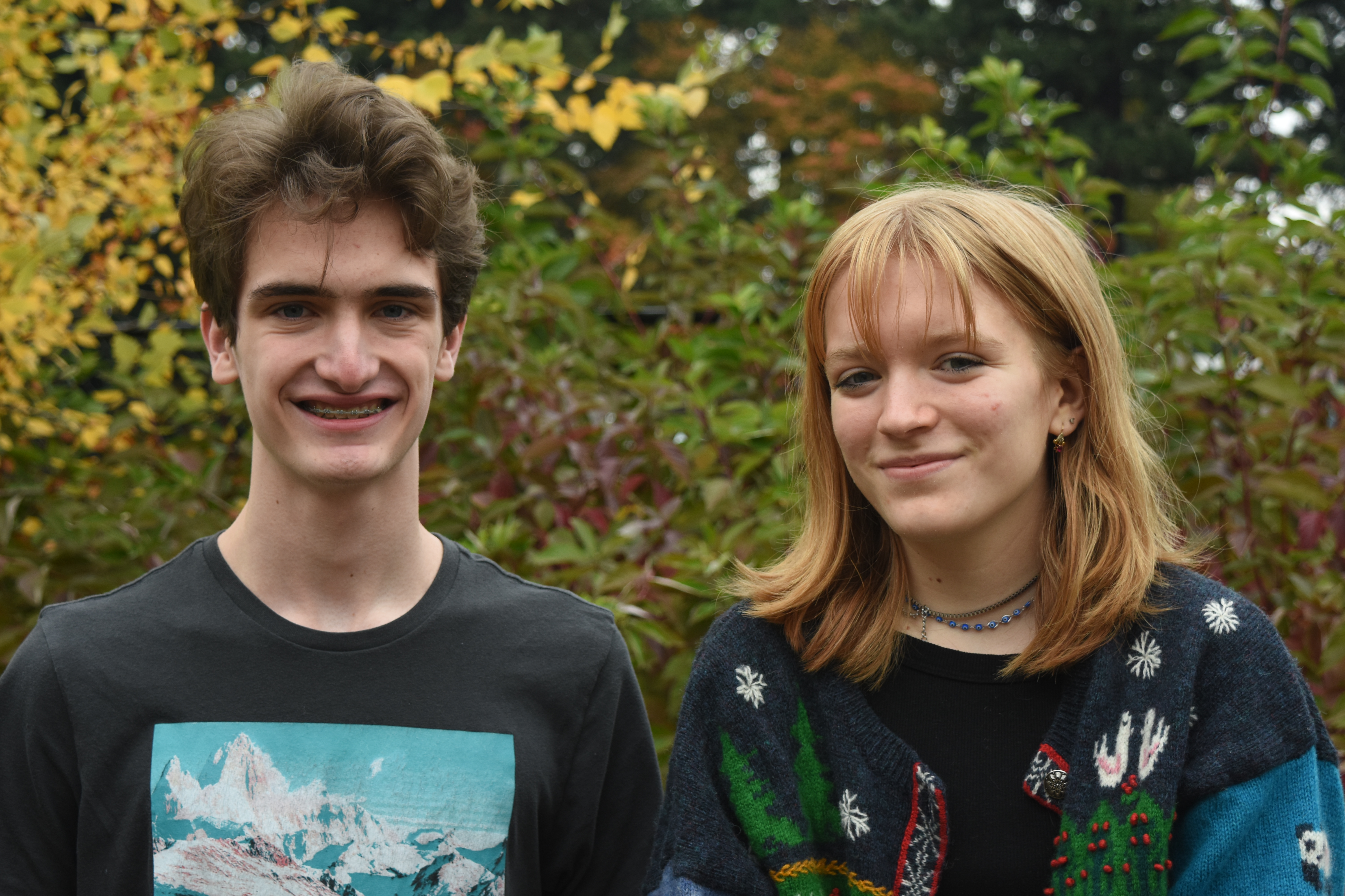 Two white teenagers, one with short brown hair and another with strawberry blonde hair and bangs, stand in front of a wall of greenery, smiling. 