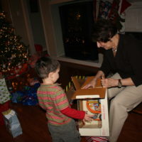 A small child stands by as their grandmother opens a toy box for them, a lit up Christmas tree ringed by presents stands in the background
