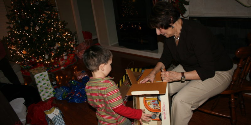 A small child stands by as their grandmother opens a toy box for them, a lit up Christmas tree ringed by presents stands in the background