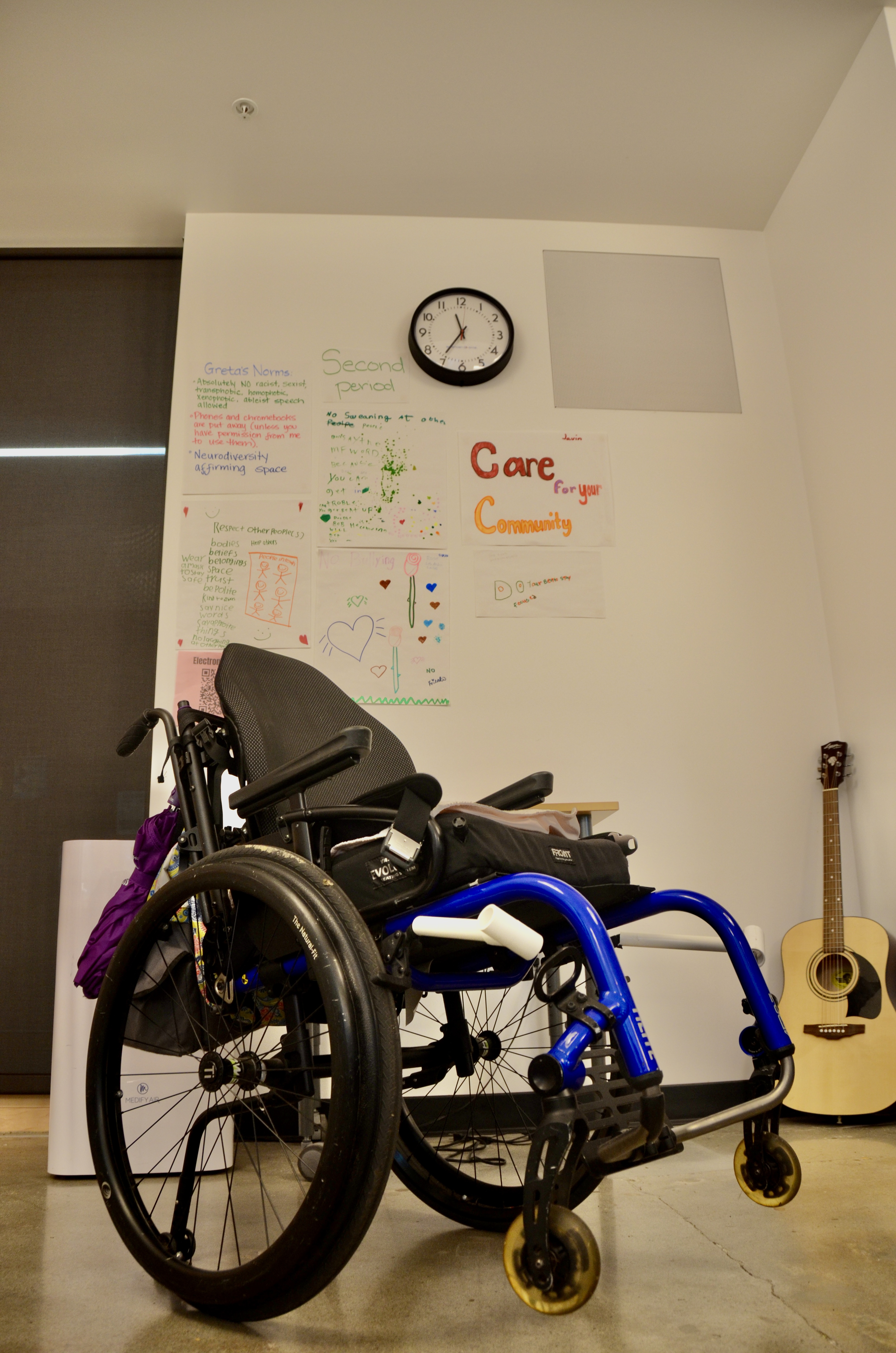 Against a white wall, a black wheelchair is set at an angle. A guitar also leans against the wall on the right side of the photo.