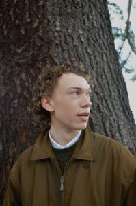 A white teenage boy wearing a brown jacket looks off to the side as he stands in front of a tree trunk