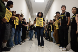 A student holding a sign saying "Green New Deal for Schools Now" stands in the middle of the hallway, surrounded by other students activists on either side.