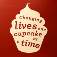 A red sign with a white image of a cupcake with the phrase, "Changing lives one cupcake at a time," in red lettering is posted in Sarah Bellum's Bakery & Workshop.