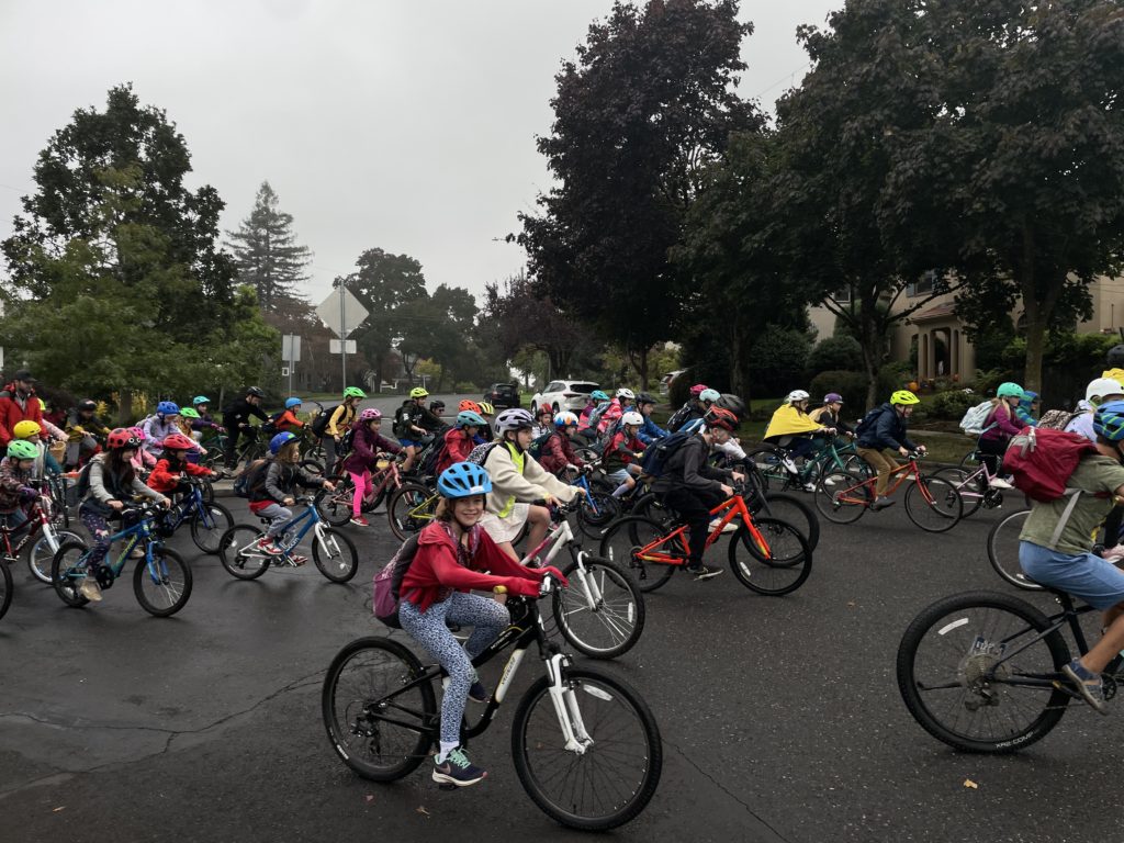 A photo of a group of elementary schoolers biking down the street on a cloudy day, with a girl at the forefront of the photo smiling at the camera while biking.