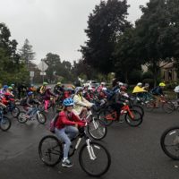 A photo of a group of elementary schoolers biking on a cloudy day, with a girl on a bike at the forefront of the photo smiling at the camera.