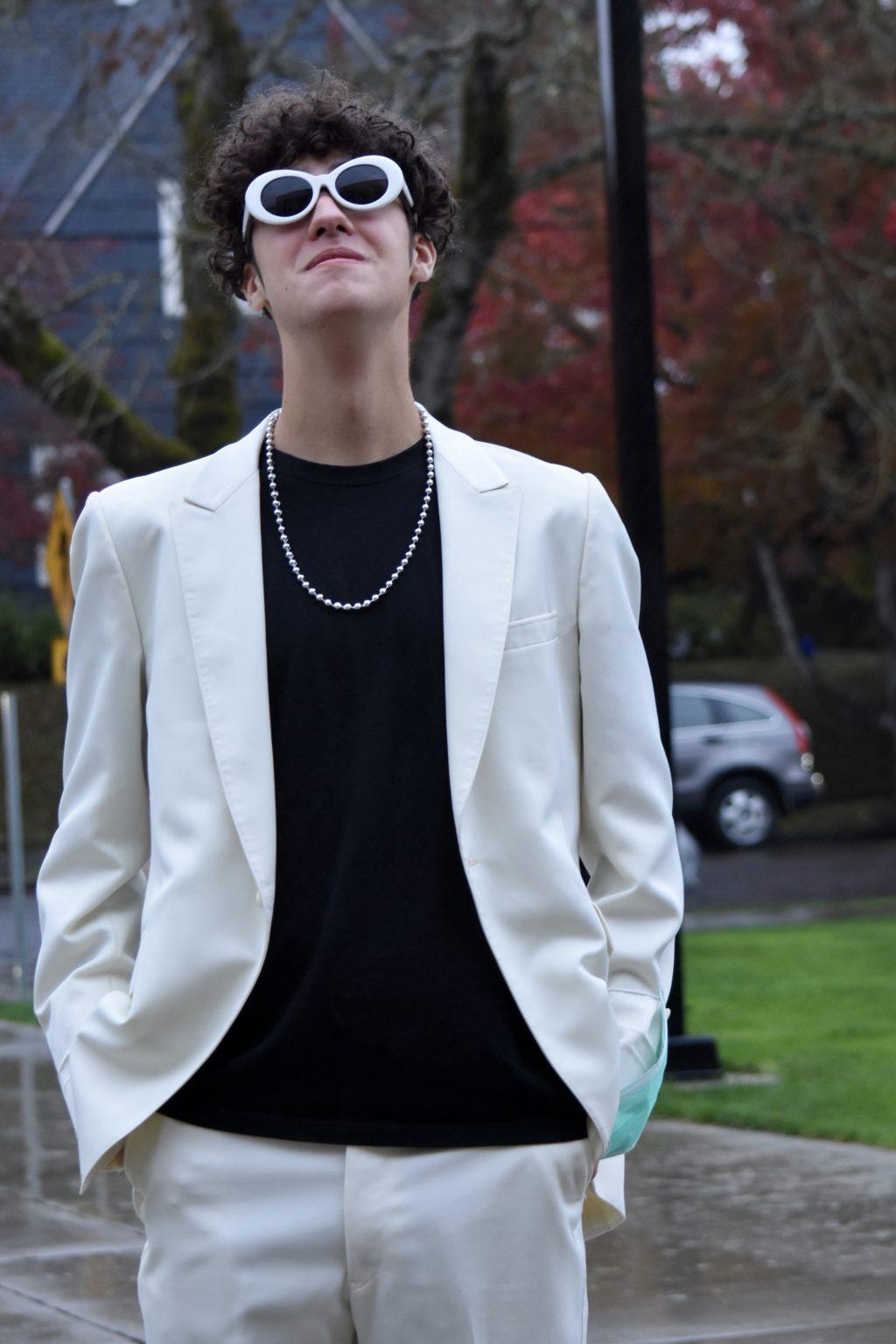 Mizani wears white pants and a white blazer over a black T-shirt. The costume is accessorized with a beaded necklace and white sunglasses. 