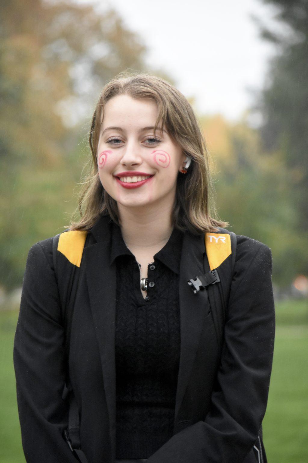 Langton stands in a black shirt and black blazer, with red swirls painted onto her cheeks. 