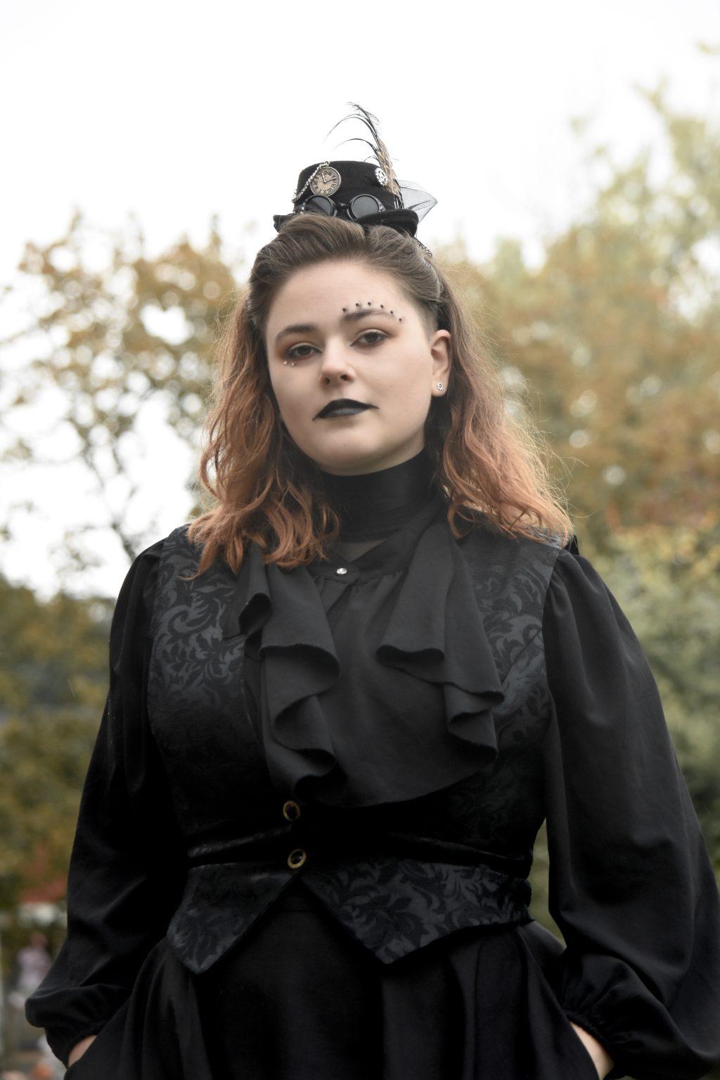 Fitzgerald wears a black vest and blouse and a tiny hat decorated with a feather and pocket watch. They have black makeup around their eyes and wear black lipstick. 