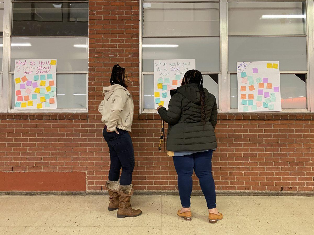 Two parents wearing fleece jackets point to a brick wall with a white poster covered in colorful sticky-notes.