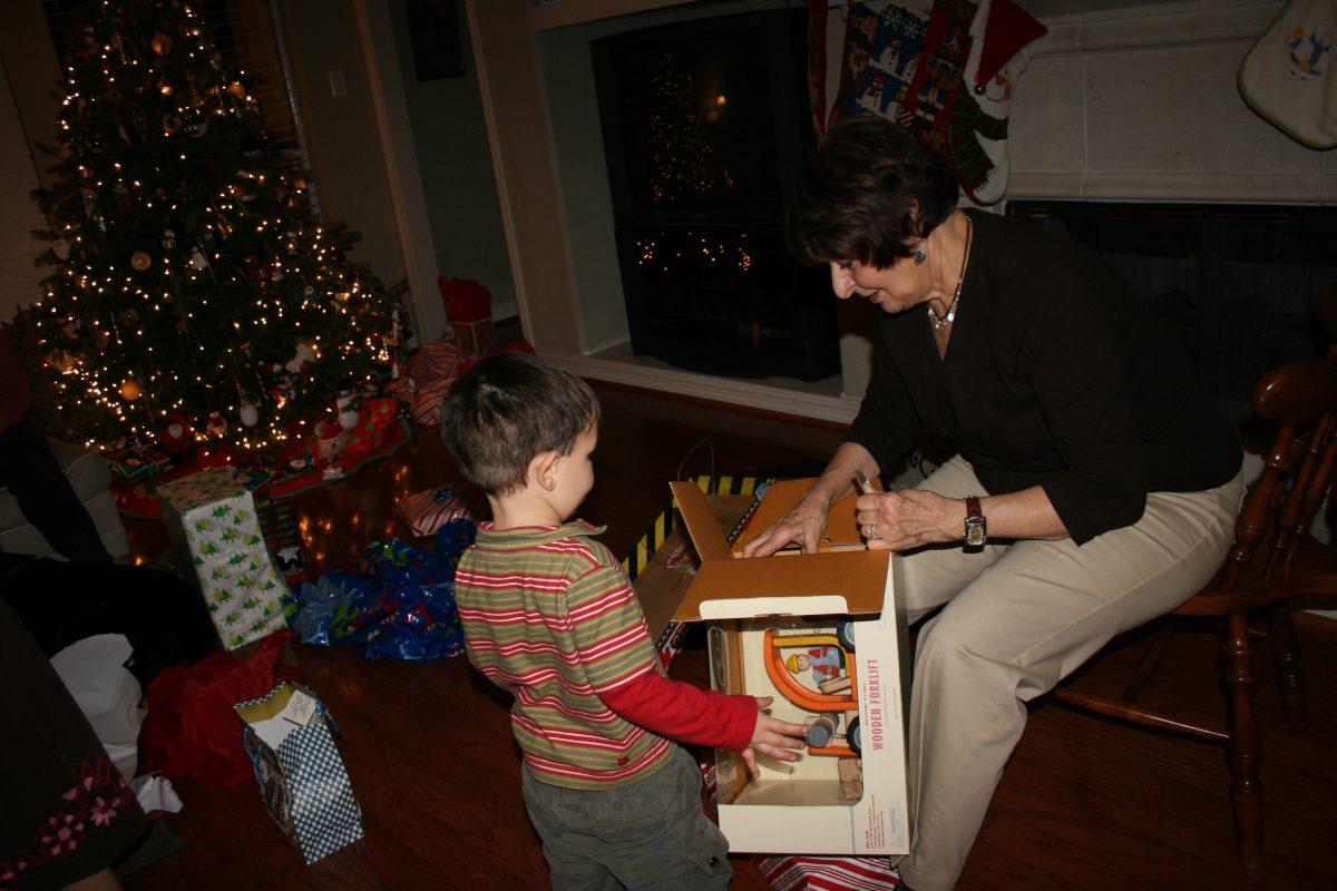 A small child stands by as their grandmother opens a toy box for them, a lit up Christmas tree ringed by presents stands in the background