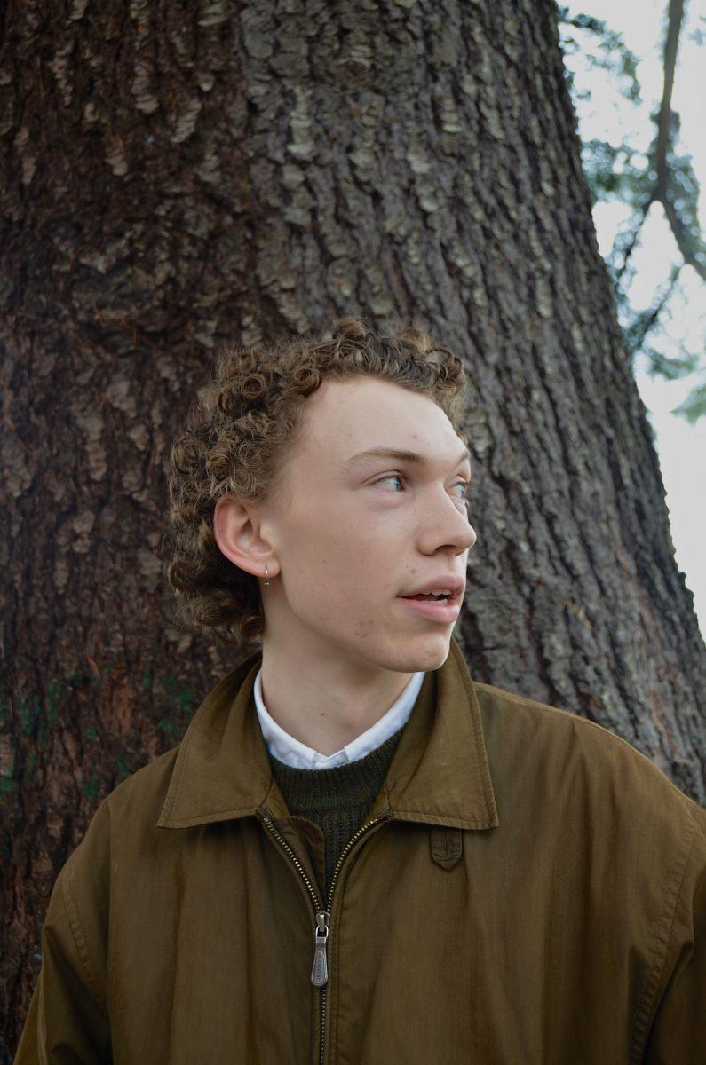 A white teenage boy wearing a brown jacket looks off to the side as he stands in front of a tree trunk