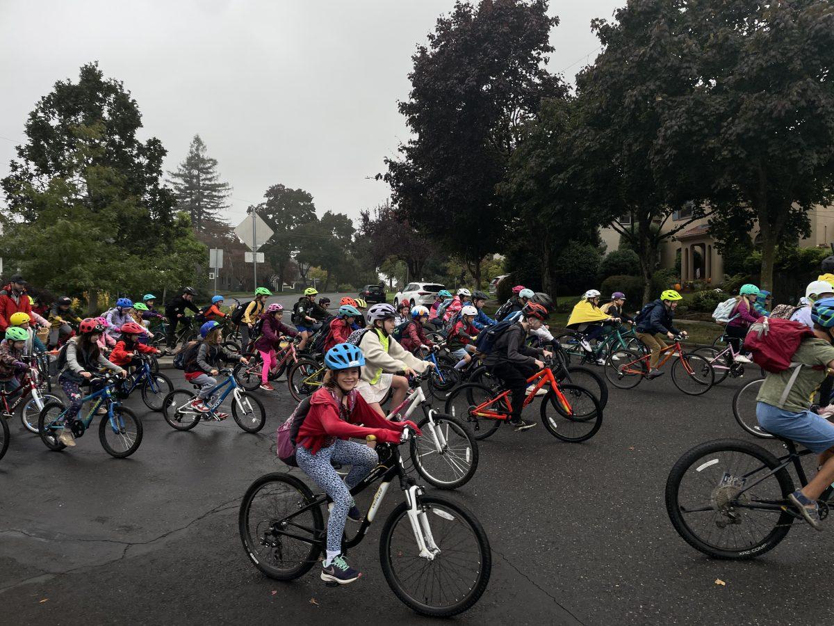 A photo of a group of elementary schoolers biking on a cloudy day, with a girl on a bike at the forefront of the photo smiling at the camera.